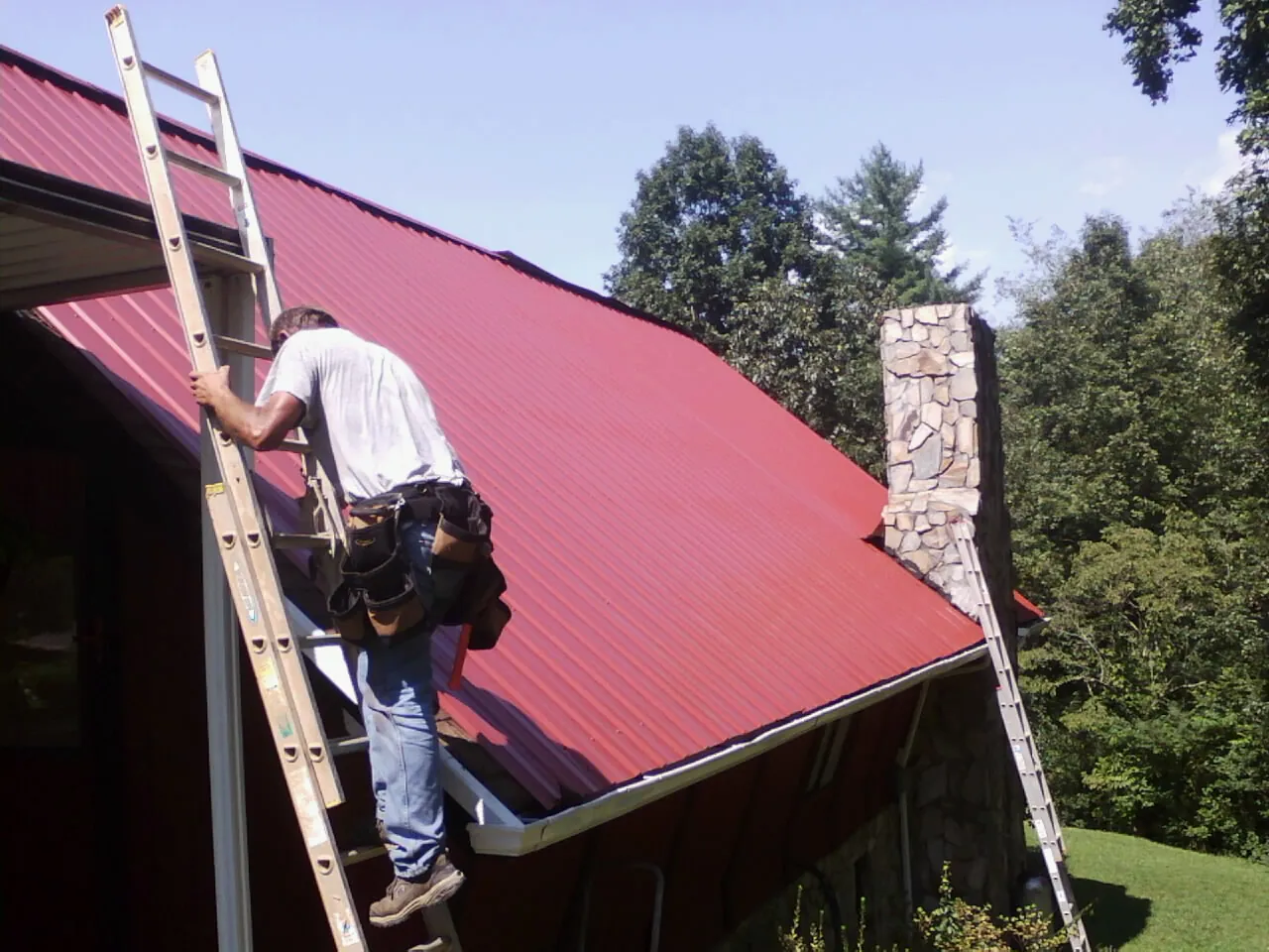 Roofer on a ladder beside a red metal roof and stone chimney — Soffit & Fascia Repair by Roofing Brothers in Dahlgren
